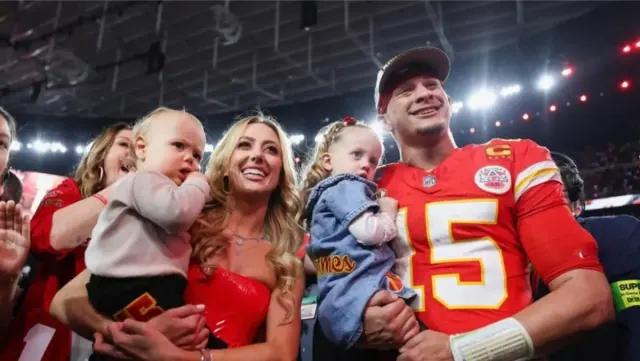 Brittany and Patrick Mahomes and their two children. They are holding their children in their arms and are looking at something and smiling.