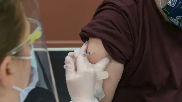 A man wearing a mask receiving a vaccine shot from a person wearing gloves, a mask, and a face shield.