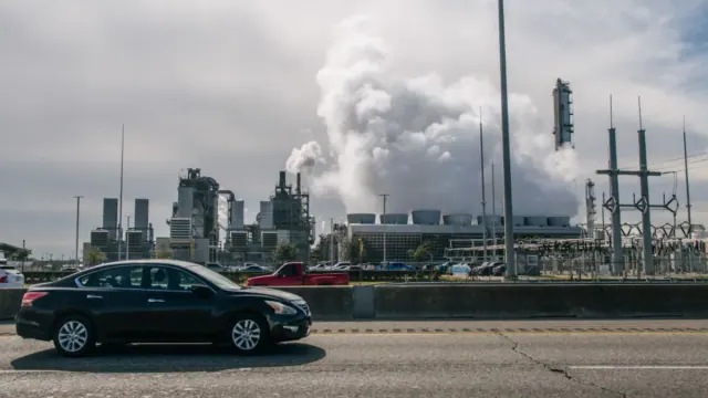 A few cars driving on a highway with a large factory in the background spewing plumes of white and grey smog