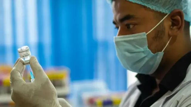 A man wearing a blue mask extracts a vaccine from a bottle using a needle.