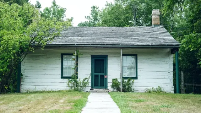 A small white house with a green door and two windows. The boards on the outside of the house are in poor repair and the lawn is mostly brown