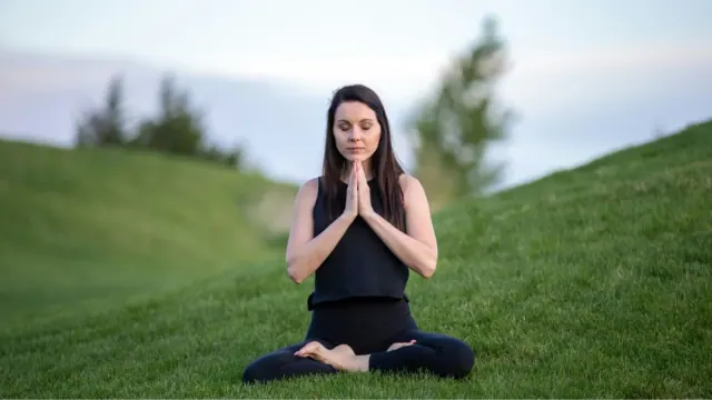 A woman sitting down on the grass outside with her hands clasped in front of her meditating.