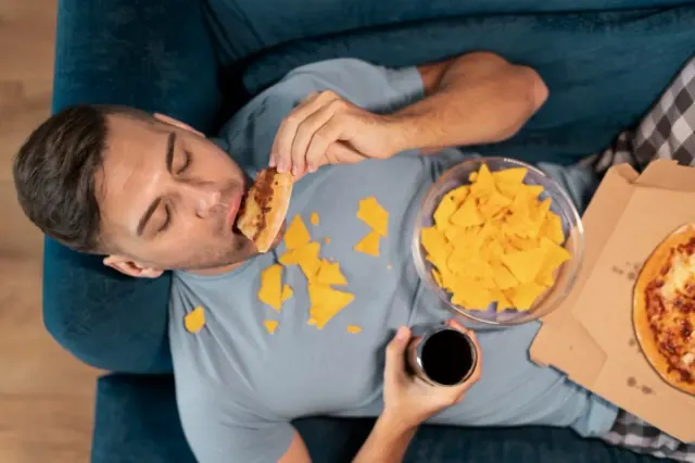 A man is pictured eating chips and a pizza on the couch