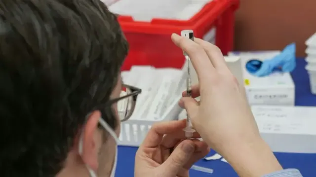 A person with glasses extracts a vaccine from a bottle by using a shot.