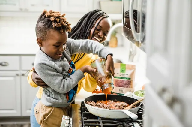 An adult beside a child pouring sauce into a pan in an archive kitchen photograph.