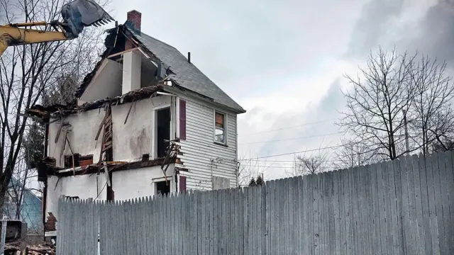 A two-story white house in disrepair is torn down with a large machine next to a grey wood fence