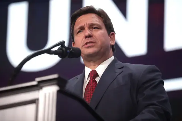 Ron DeSantis in a black suit and red tie standing on a stage addressing a crowd through a microphone