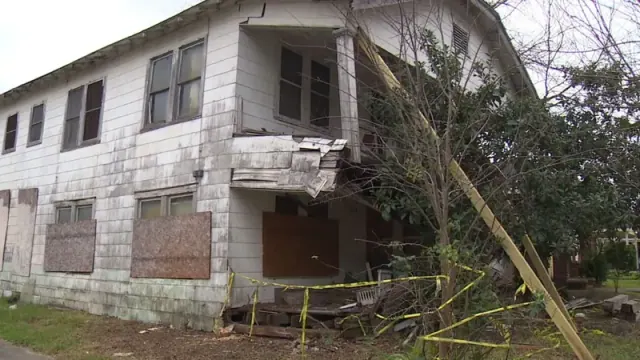 A white house in terrible condition with boards on the window and overgrown trees in the front yard