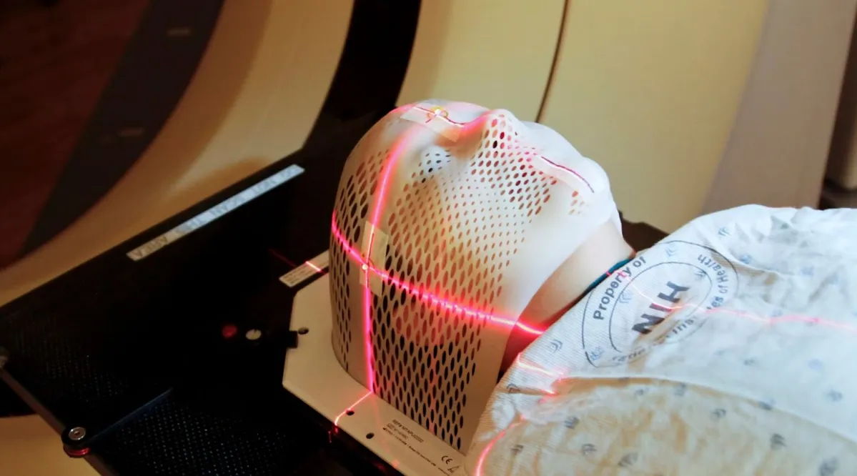 A person lying down with a mesh mask over their head about to go through a scanner. There are red lights on the mask.