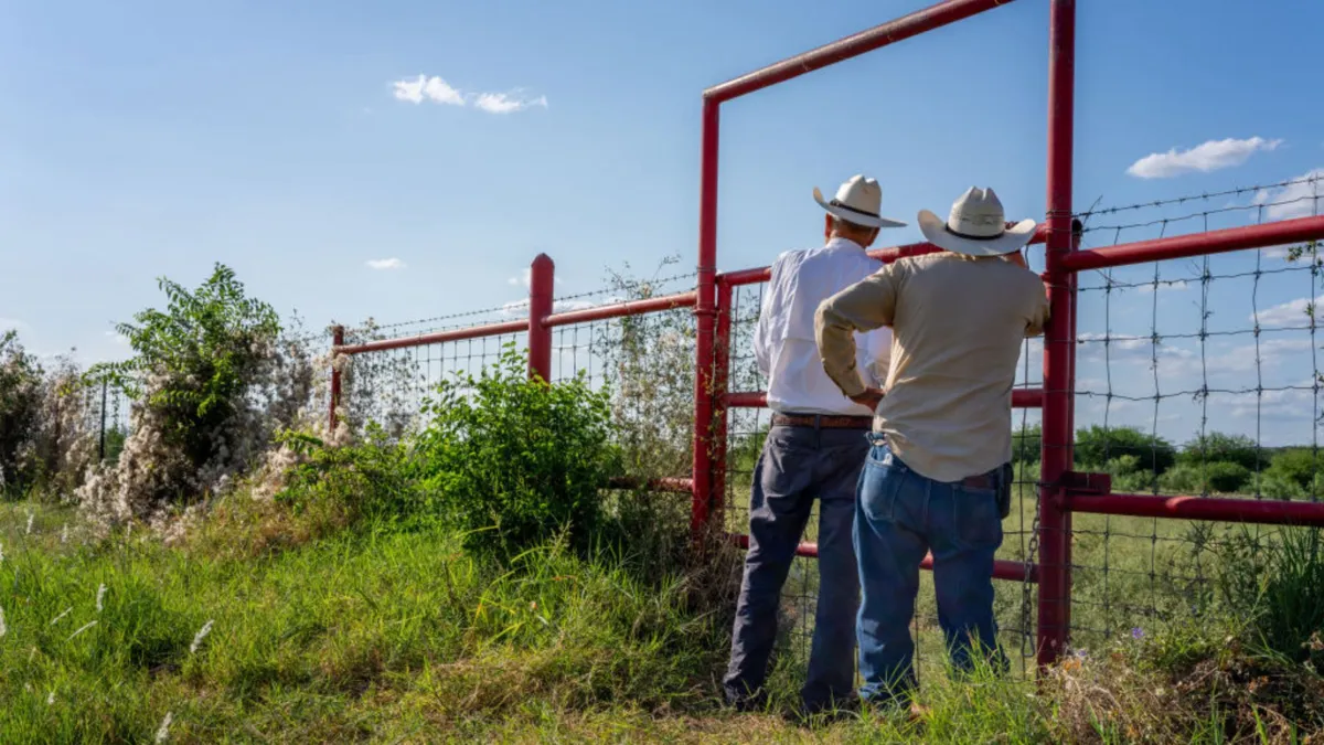 Two ranchers wearing jeans, button down shirts, and hats stand against a red fence surrounded by greenery