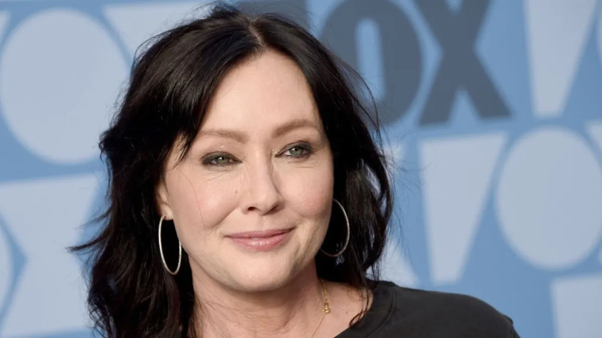 Shannen Doherty, smiling, with shoulder-length black hair, wearing hoop earrings and a dark top, poses for the camera at a public event with a blue and white geometric patterned backdrop