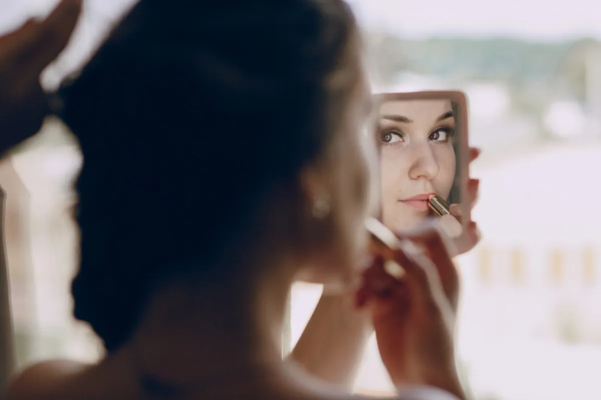 A woman is pictured looking in a mirror as she applies lipstick