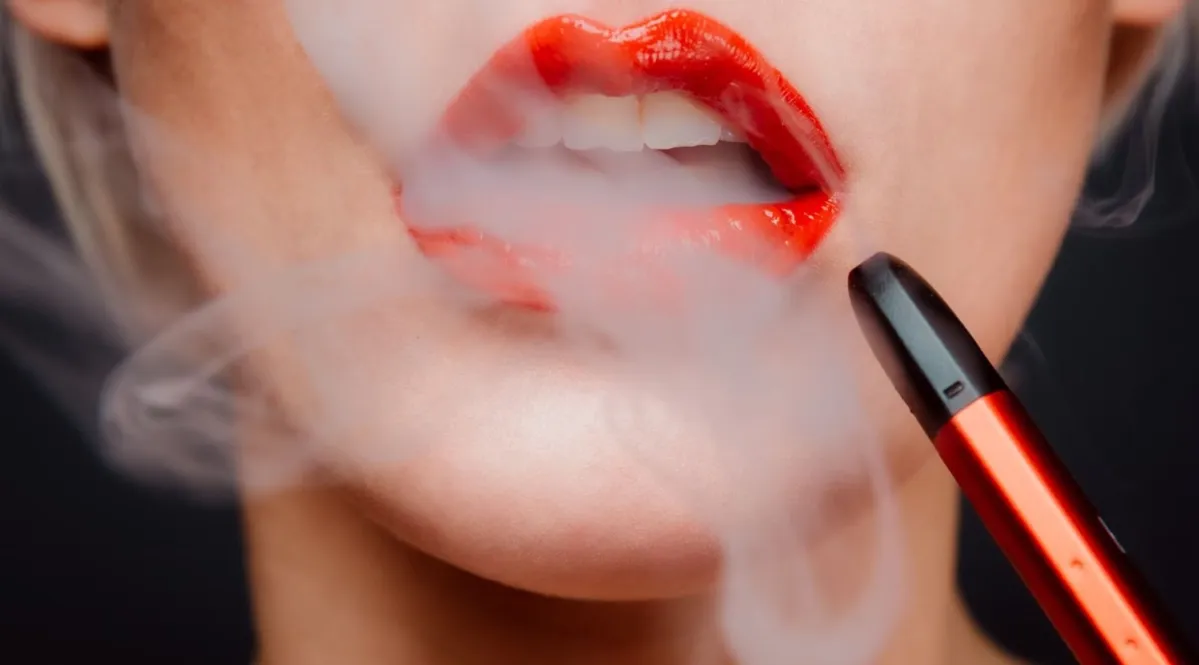 A woman wearing red lipstick and vaping with a red and black vape.