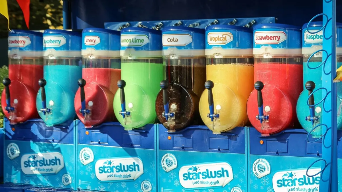 A row of colorful iced slushy machines labeled with different flavors such as strawberry, cherry, lemon lime, cola, tropical, and raspberry. The machines have clear containers showcasing the brightly colored slushy drinks