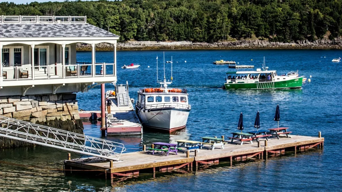 A waterfront scene showing a large grey house with a covered porch built over a rocky shore. A metal gangway leads down to a wooden dock with purple and green picnic tables. Two boats are in the water