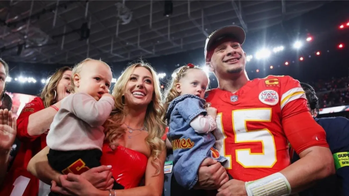 Brittany and Patrick Mahomes and their two children. They are holding their children in their arms and are looking at something and smiling.