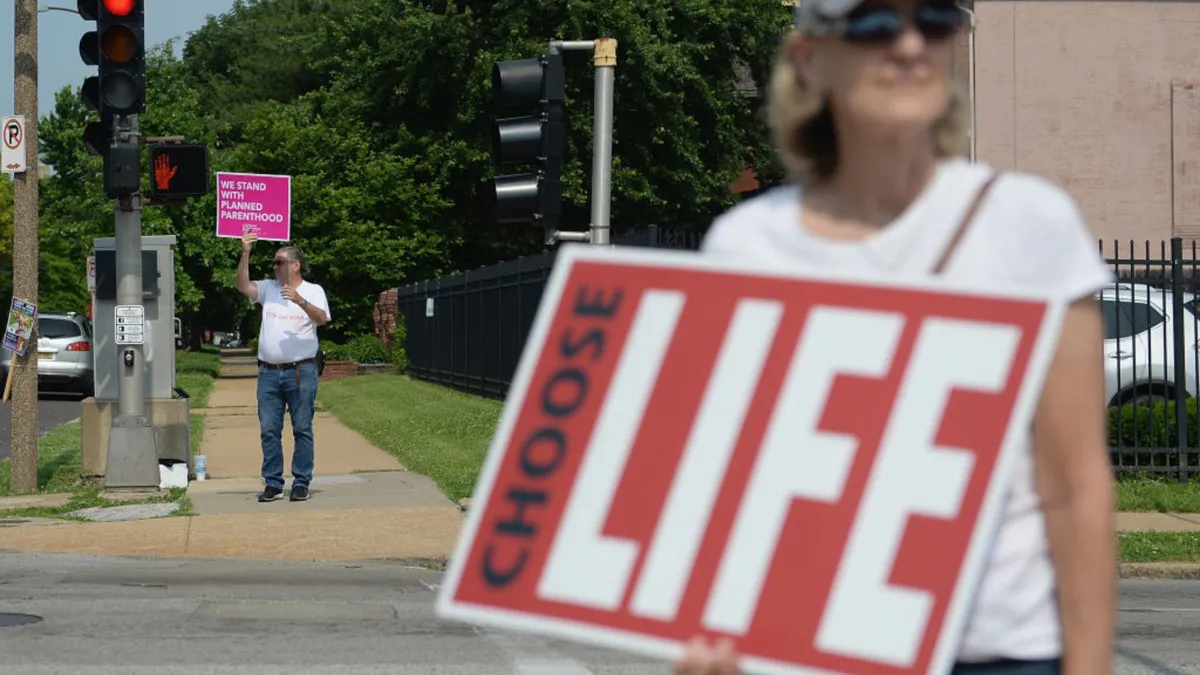 A woman wearing a shite t shirt holding a sign that reads "CHOOSE LIFE" with another man in the background holding a pink sign