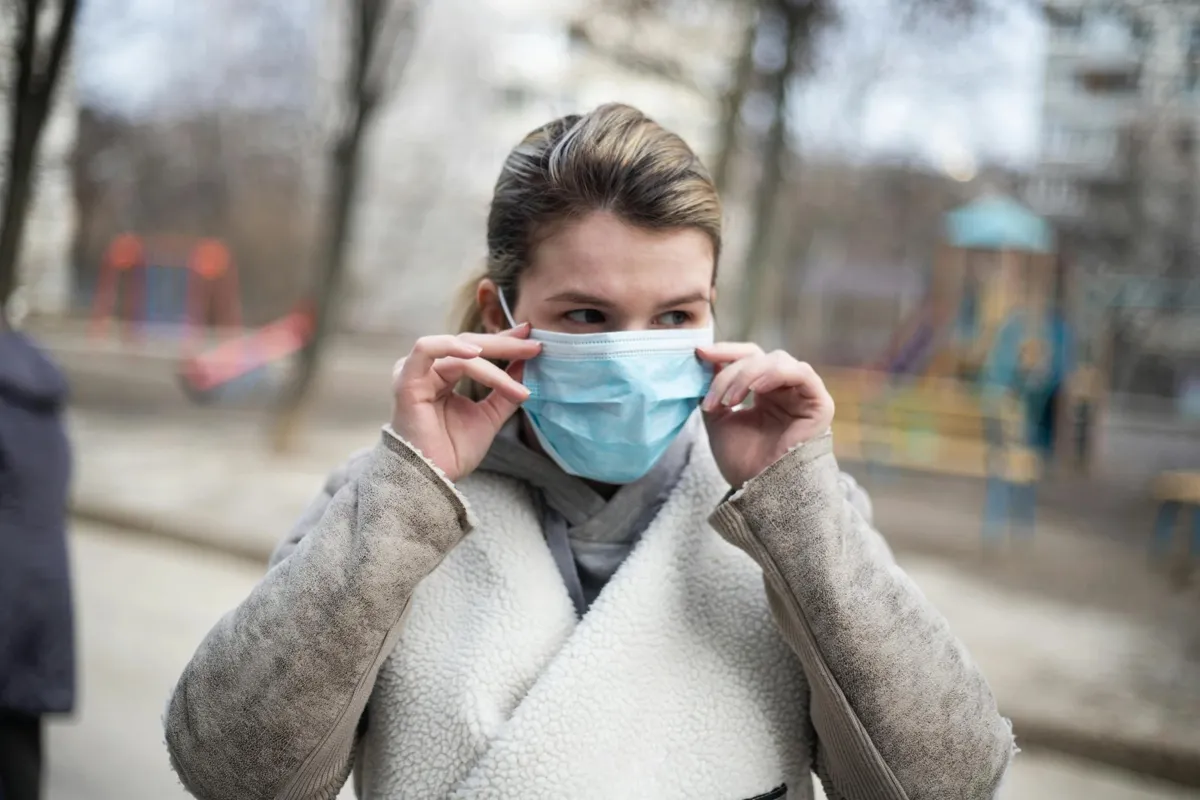 A woman with blonde hair in a ponytail putting. a mask on as she walks outside