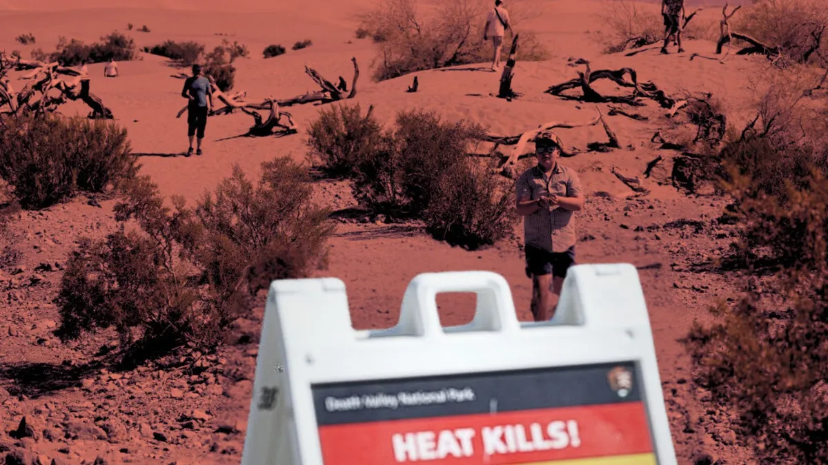 A sandwich board sign with a warning that says "heat kills!" with people walking around in a desert setting behind