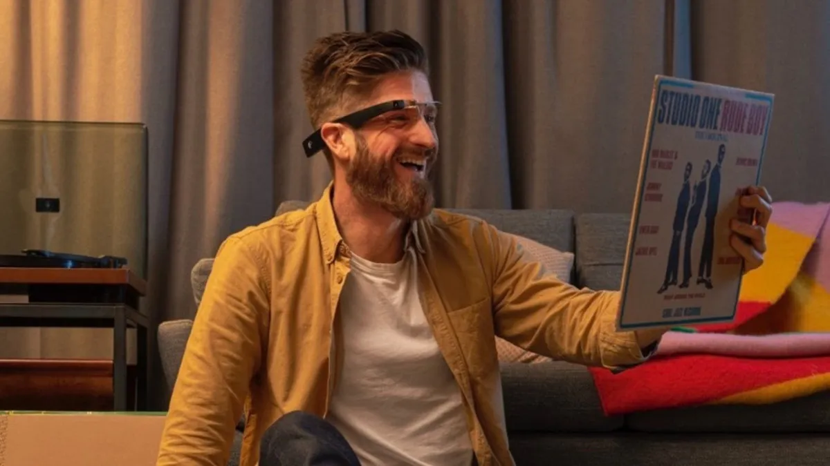 A smiling man with a beard, wearing smart glasses and a yellow shirt under an unbuttoned jacket, holds up a vinyl record cover