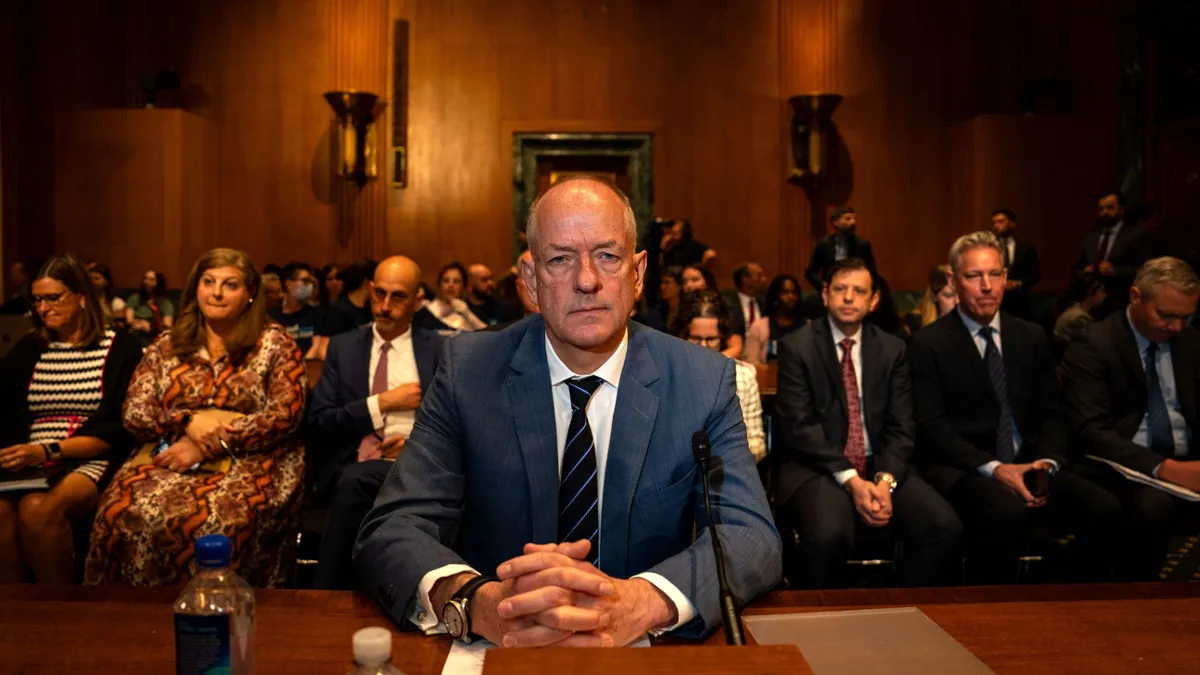 A bald man wearing a blue suit sits at a large wood table with his hands crossed with many people sitting in benches behind him