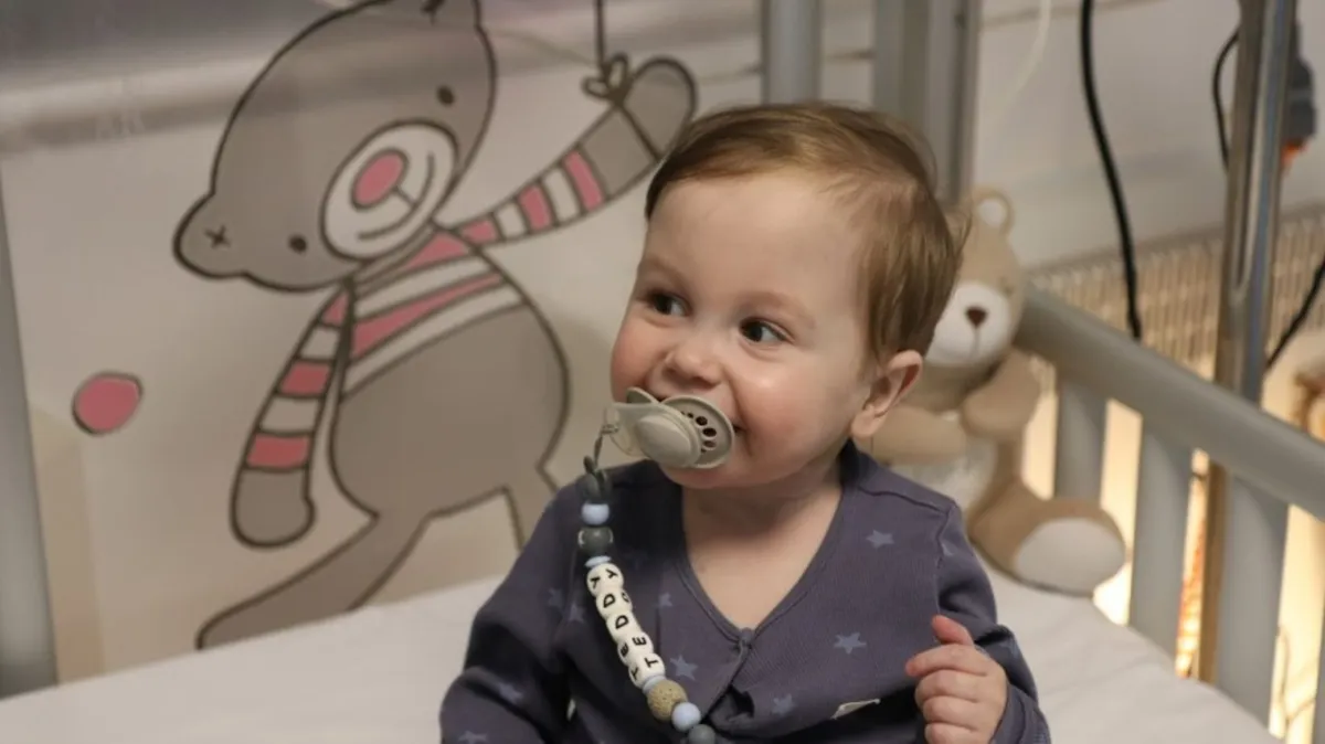 A smiling toddler, sitting in a hospital crib. The child is wearing a blue onesie with white stars and has a pacifier attached to a pacifier clip with beads spelling out the name "TEDDY.