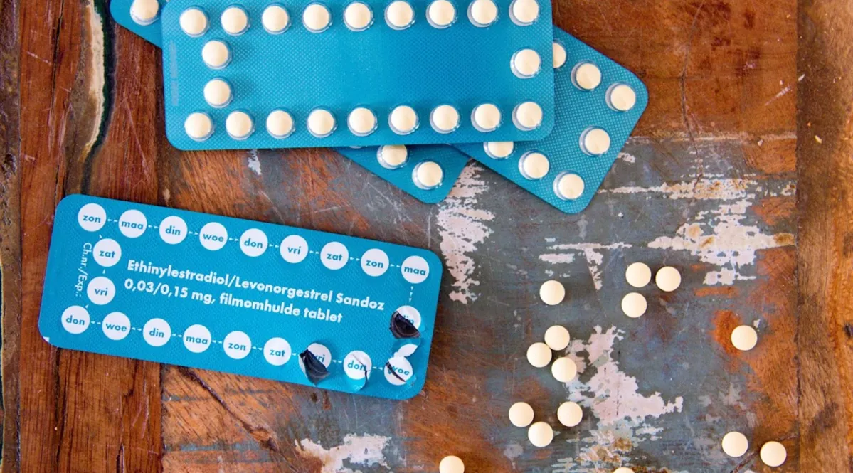 Four packets of blue birth control pills on a wooden table. Three packets are perfectly intact, whereas one of them has been opened. There are a few pills that have been scattered across the table.