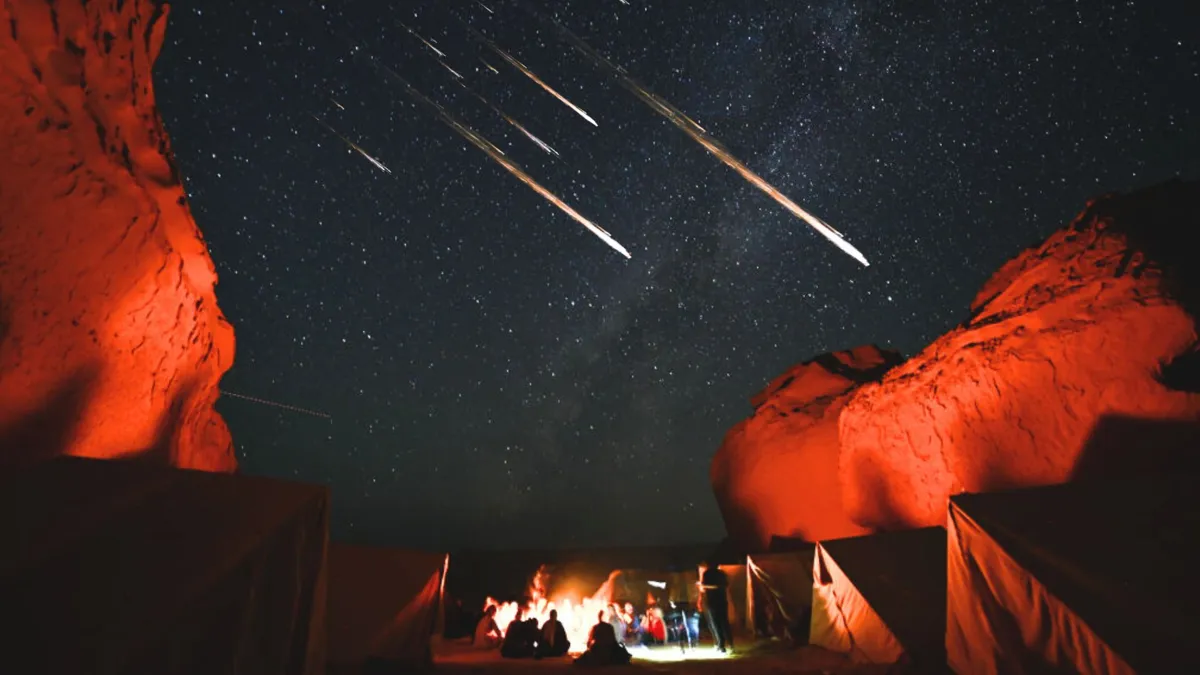 A group of people camping next to illuminated red rocks under a large meteor shower