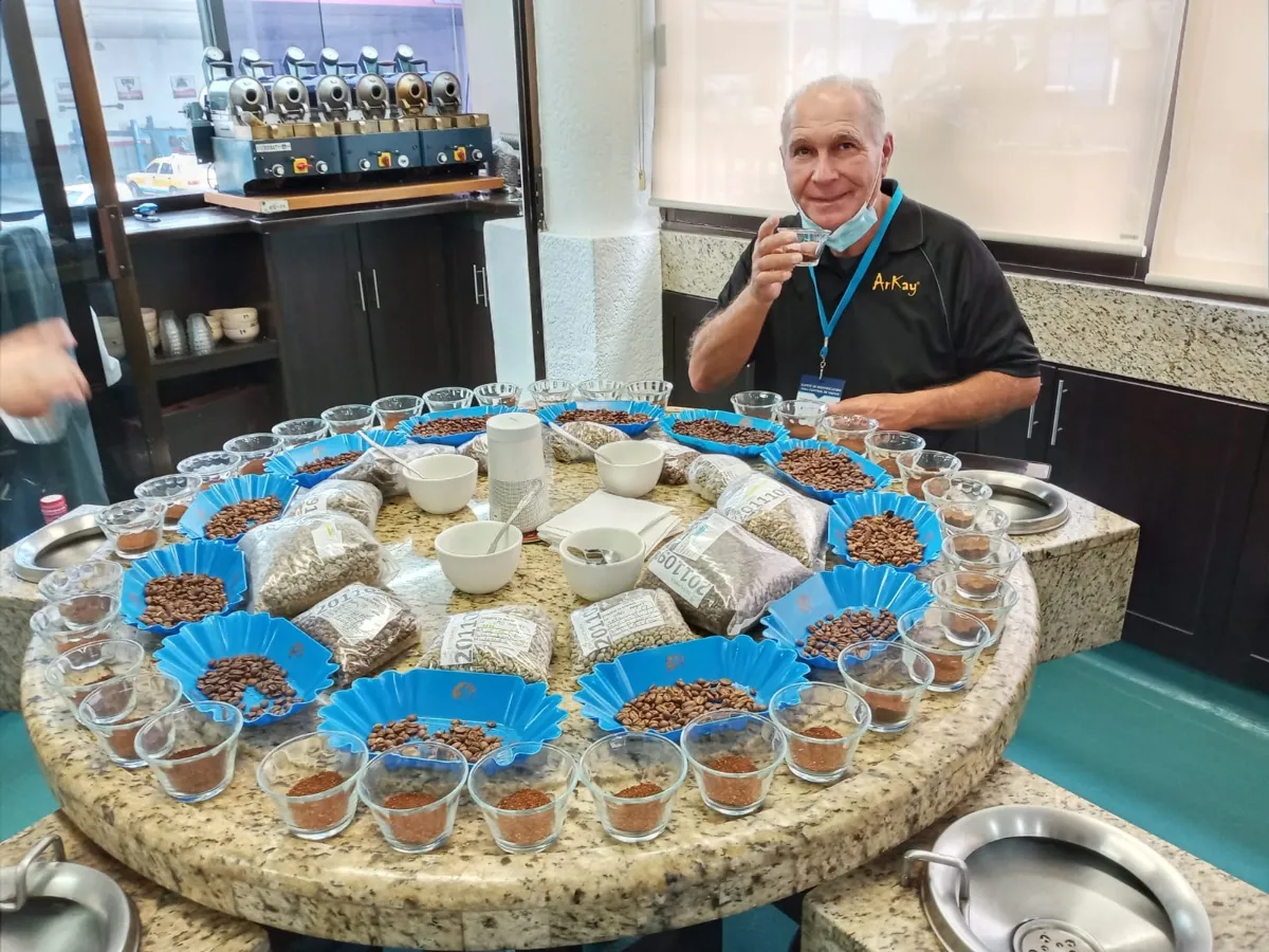 A man sits at a round table where there are cups of coffee beans. He's wearing a face mask and sampling coffee