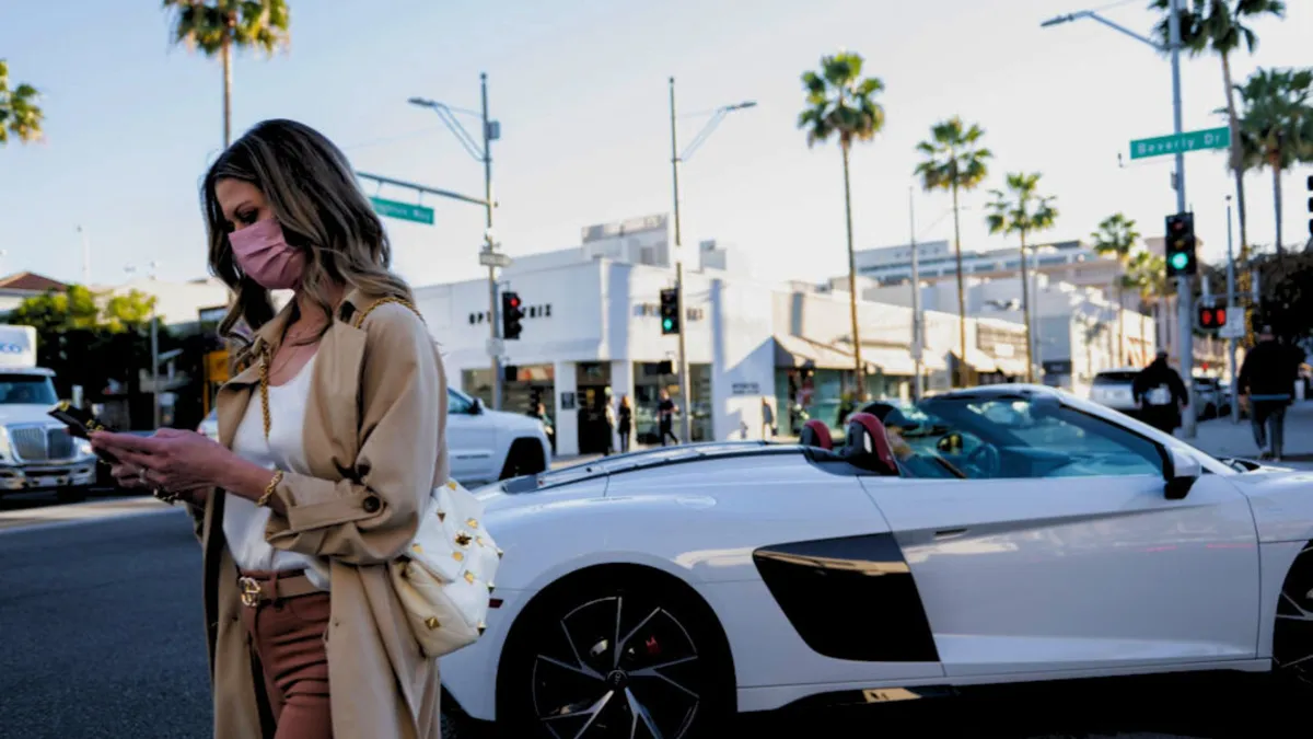 A woman wearing a trench coat and a mask walks past a sports car on a busy downtown street with palm trees