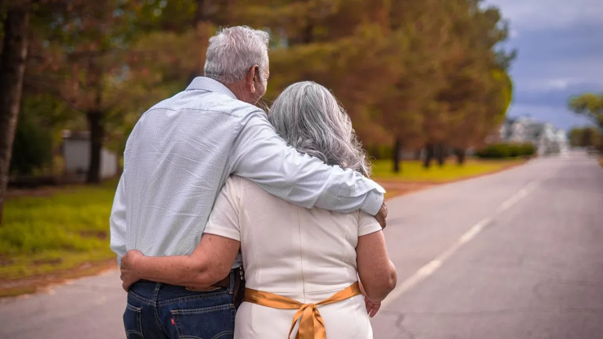An elderly couple is seen from behind, walking arm-in-arm down a deserted suburban road lined with trees