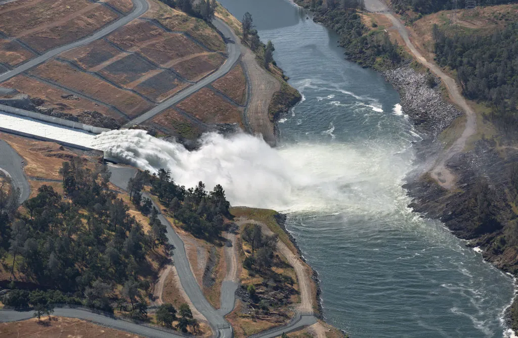 A large dam spills water into a nearby river surrounded by dry land and black roads