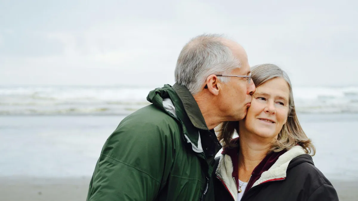 An elderly man kissing the cheek of an elderly woman in front of the ocean.