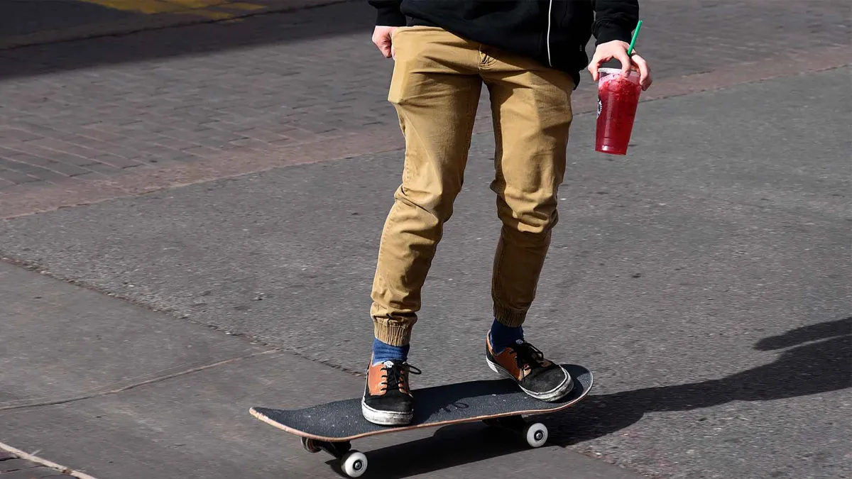 A teenager rides his skateboard along a street holding Starbucks drink