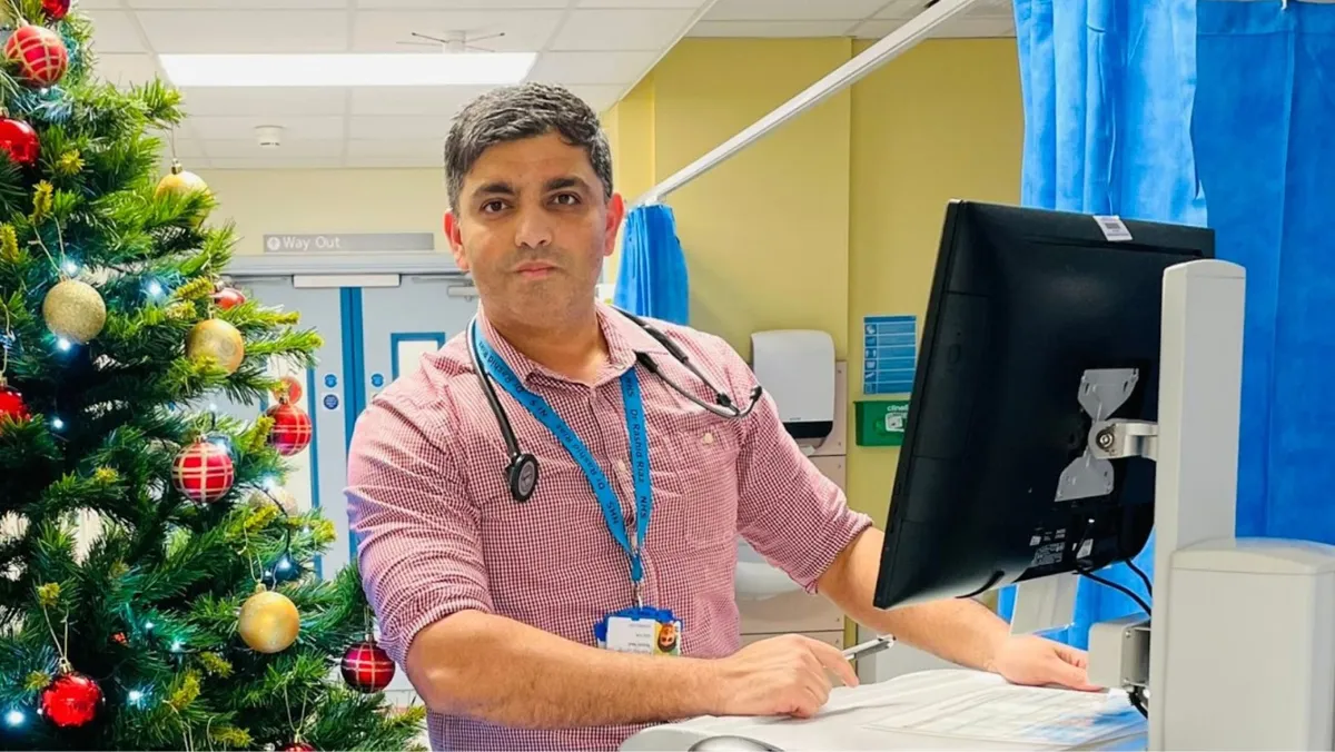 A man in a pink checkered shirt and a stethoscope around his neck stands at a hospital workstation. Behind him is a decorated Christmas tree