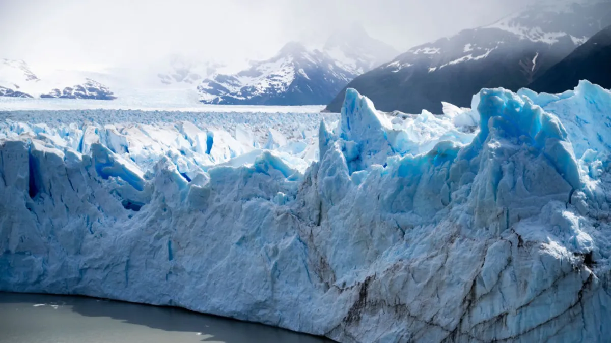 A large blue and white glacier next to the ocean in front of large snow capped mountains