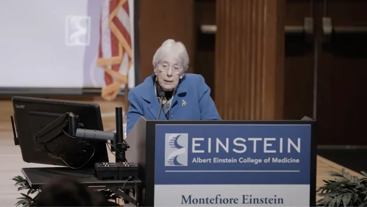 An elderly woman wearing a blue blazer and glasses is speaking at a lectern with the Albert Einstein College of Medicine and Montefiore Einstein logos displayed
