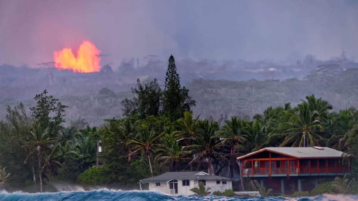 A large plume of lava erupts from a mountain behind a small city with houses and tropical trees next to the ocean