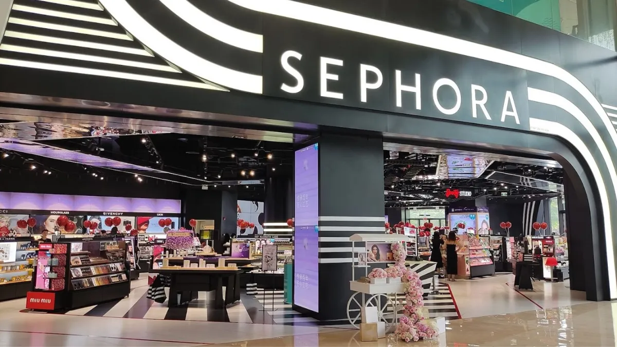 The entrance of a Sephora cosmetics store featuring a bold, black facade with the brand's name in white capital letters. Inside, the spacious store is filled with various makeup and skincare product displays, promotional banners, and a few customers browsing