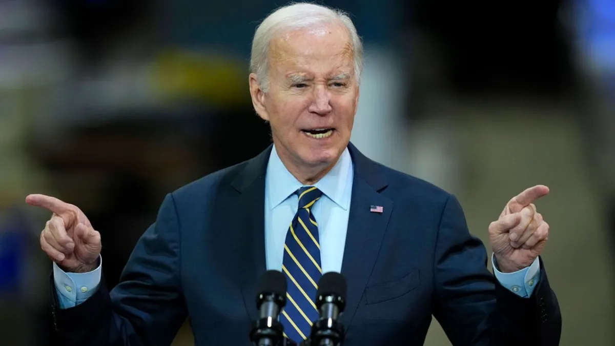 President Joe Biden stands while giving a speech and pointing his fingers at the audience. He wears a blue suit jacket, blue shirt, and striped tie
