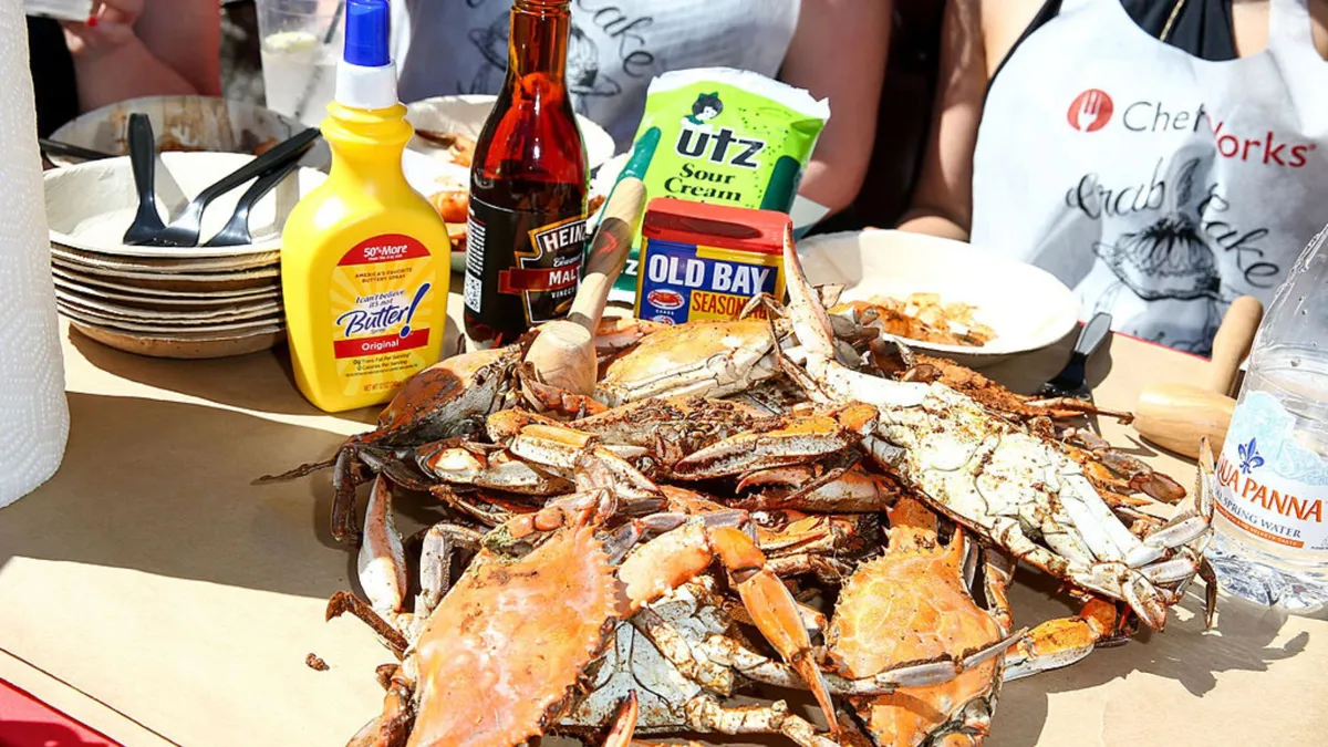 A pile of crabs on a sheet of brown paper next to a series of seasonings, people wearing crab bibs, and paper plates