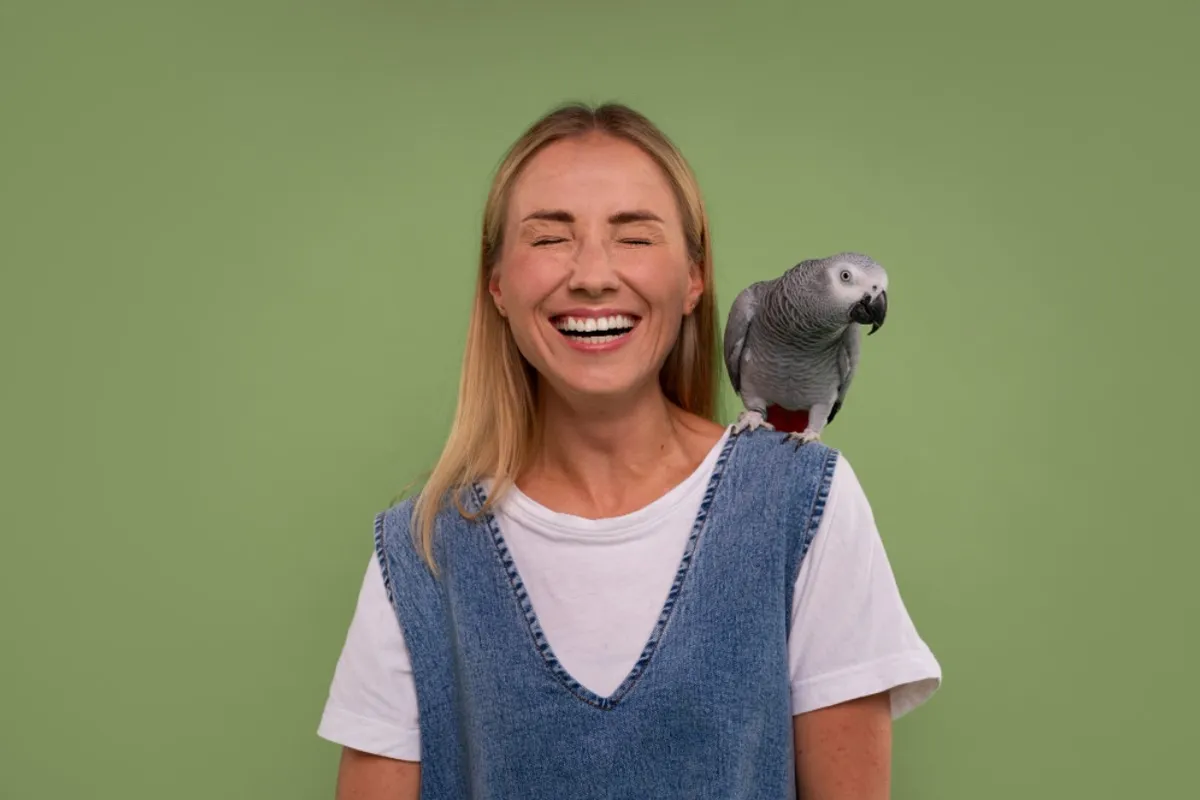 A woman dressed in denim overalls smiles with a parrot on her shoulder