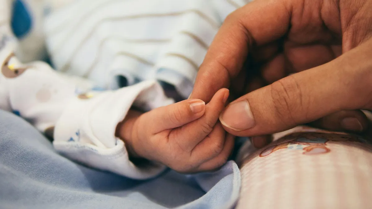 A close-up image showing a small infant's hand with tiny fingers wrapped around an adult's larger finger