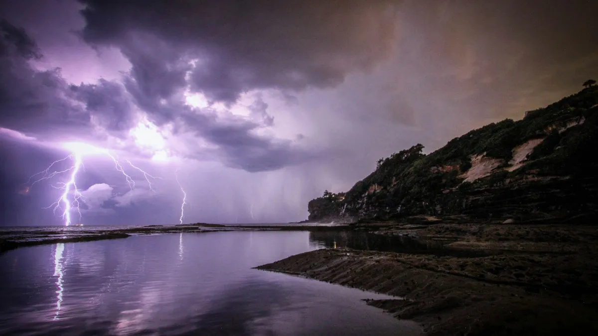 Underneath a purple sky, lightning strikes the ocean. Next to the ocean sits a sandy beach and houses on top of a cliff