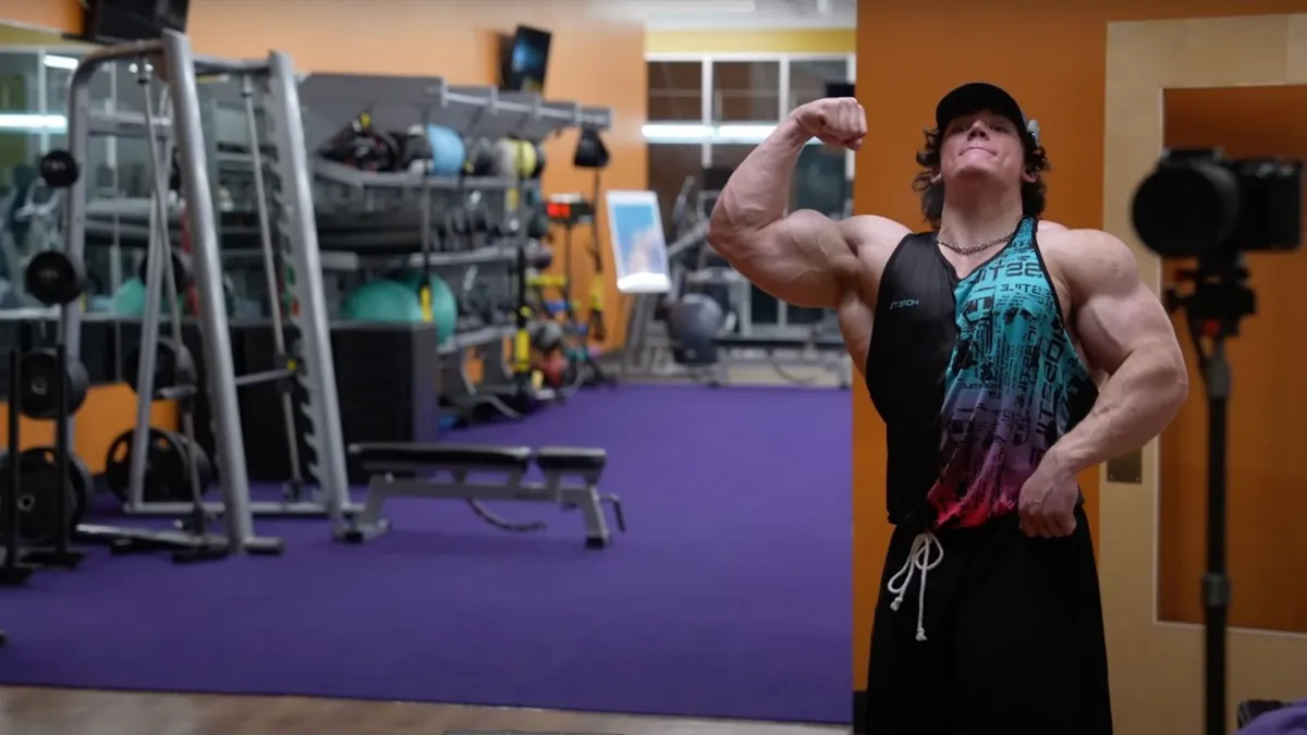 A muscular bodybuilder wearing a tank top and a baseball cap flexes his biceps while looking upward in a gym with exercise equipment in the background