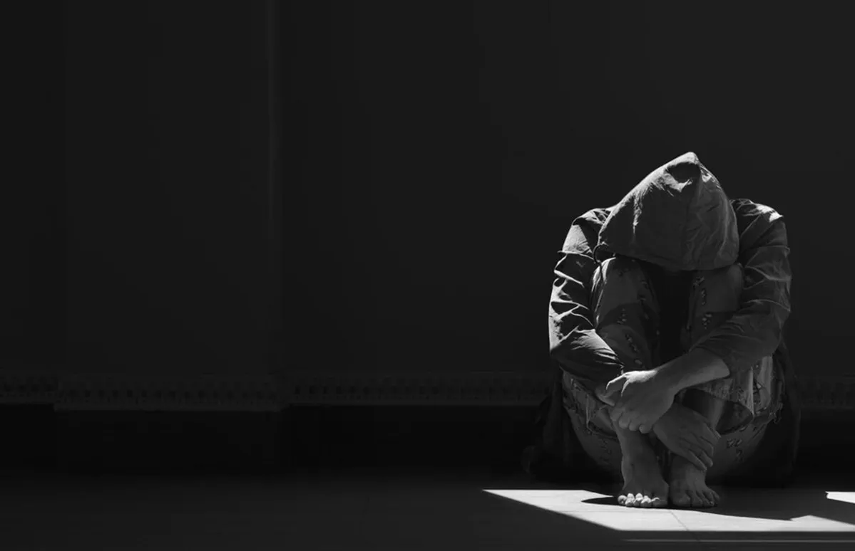 A person sits on the ground of a dark room, looking distressed