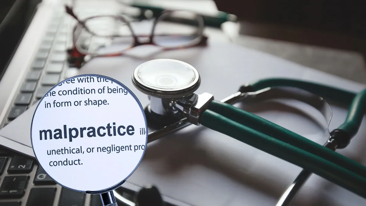 A magnifying glass hovering over the word malpractice with a stethoscope, key board, paper, and eye glasses in the background