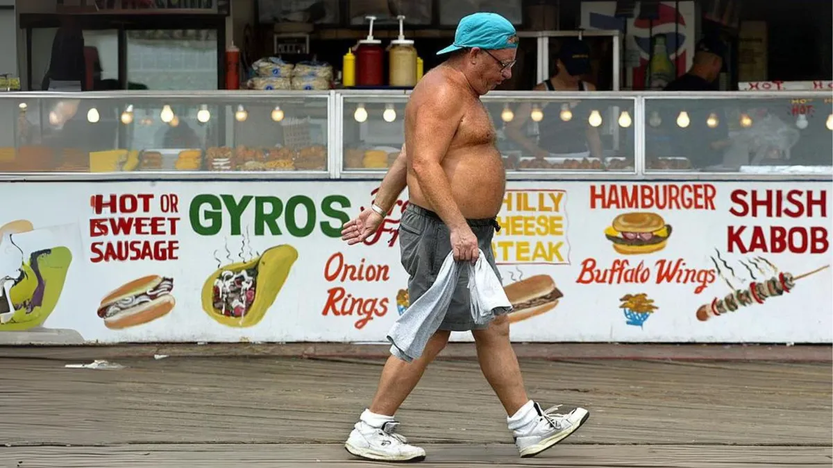 A man walking on a board walk with his shirt off in front of a sign displaying hot dogs, hamburgers, gyros and other fast food items
