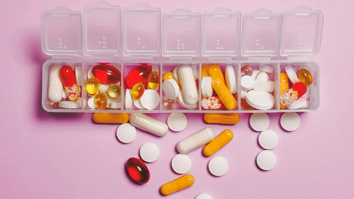 An open daily pill organizer with compartments for each day, filled with an assortment of colorful pills, and capsules on a bright pink background