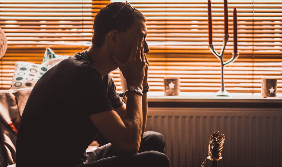 A person seated beside window blinds with a hand against their face